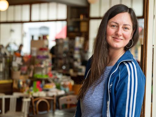 Nina Fuchs steht im Türrahmen eines Geschäfts und lächelt, sie trägt eine blaue Adidas-Jacke.