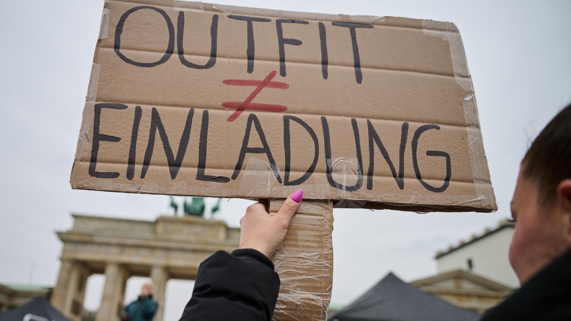 Das Bild zeigt ein Protestschild, das von einer Frau hochgehalten wird. Auf dem Schild steht "Outfit ungleich Einladung". Im Hintergrund ist das Brandenburger Tor zu erkenn. Das Bild zeigt ein Protestschild, das von einer Frau hochgehalten wird. Auf dem Schild steht "Outfit ungleich Einladung". Im Hintergrund ist das Brandenburger Tor zu erkenn.