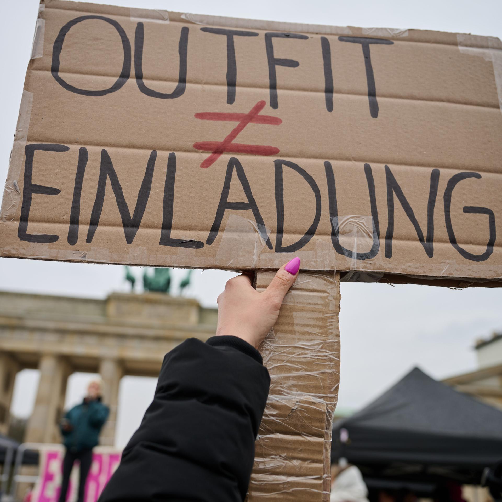 Das Bild zeigt ein Protestschild, das von einer Frau hochgehalten wird. Auf dem Schild steht "Outfit ungleich Einladung". Im Hintergrund ist das Brandenburger Tor zu erkenn.