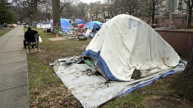 Mehrere Zelte stehen auf einem Stück Wiese. Dahinter sind Häuser zu sehen. Vor den Zelten steht eine Park-Bank. Darauf sitzt ein Mann.