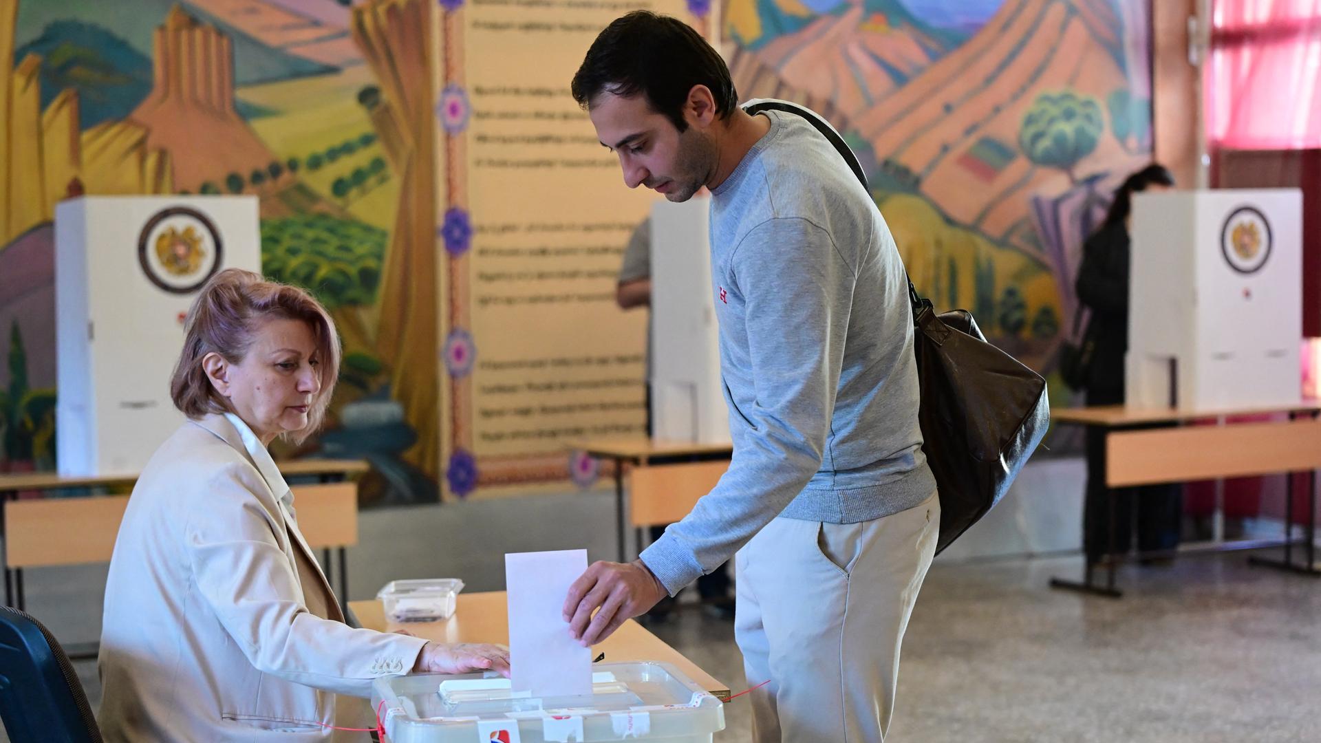 Ein dunkelhaariger junger Mann wirft einen Stimmzettel in eine Wahlurne. Eine Wahlhelferin sitzt am Tisch. 