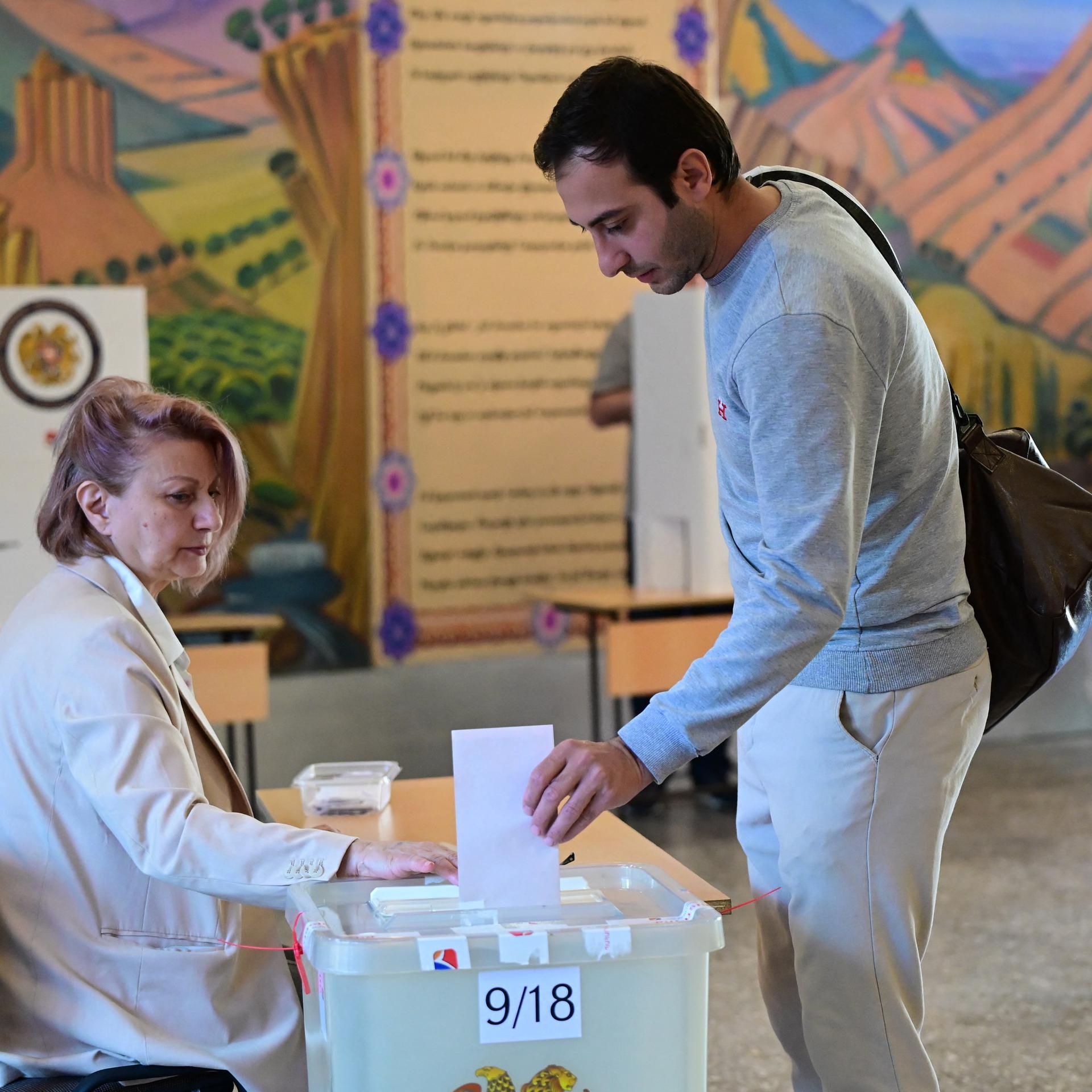 Ein dunkelhaariger junger Mann wirft einen Stimmzettel in eine Wahlurne. Eine Wahlhelferin sitzt am Tisch. 