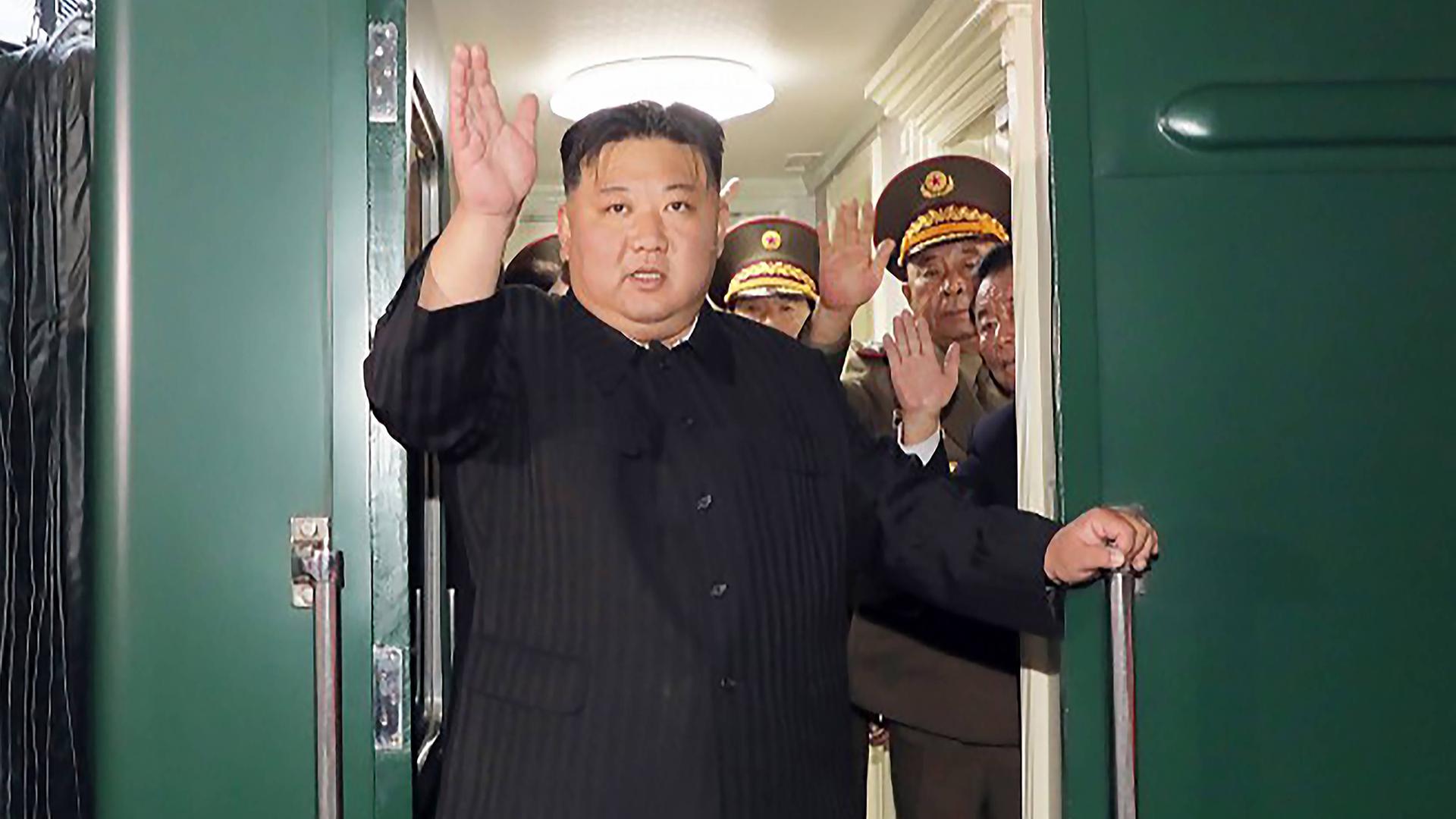 Kim Jong Un steht in schwarzem Anzug winkend in einer Zugtür.
