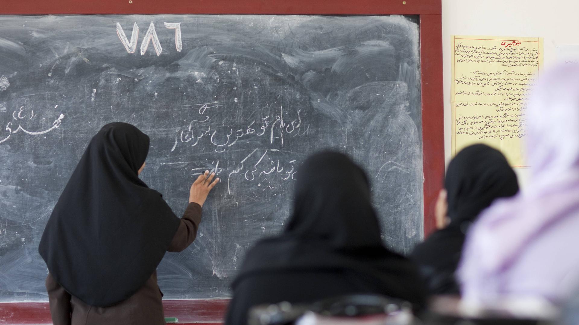 Vier Frauen im Kopftuch vor einer Tafel, den Rücken den Betrachtern zugewandt. Eine schreibt in arabischen Schriftzeichen. 