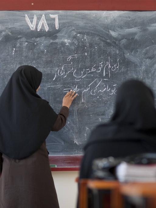 Vier Frauen im Kopftuch vor einer Tafel, den Rücken den Betrachtern zugewandt. Eine schreibt in arabischen Schriftzeichen. 