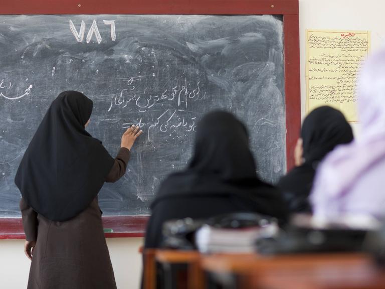 Vier Frauen im Kopftuch vor einer Tafel, den Rücken den Betrachtern zugewandt. Eine schreibt in arabischen Schriftzeichen. 