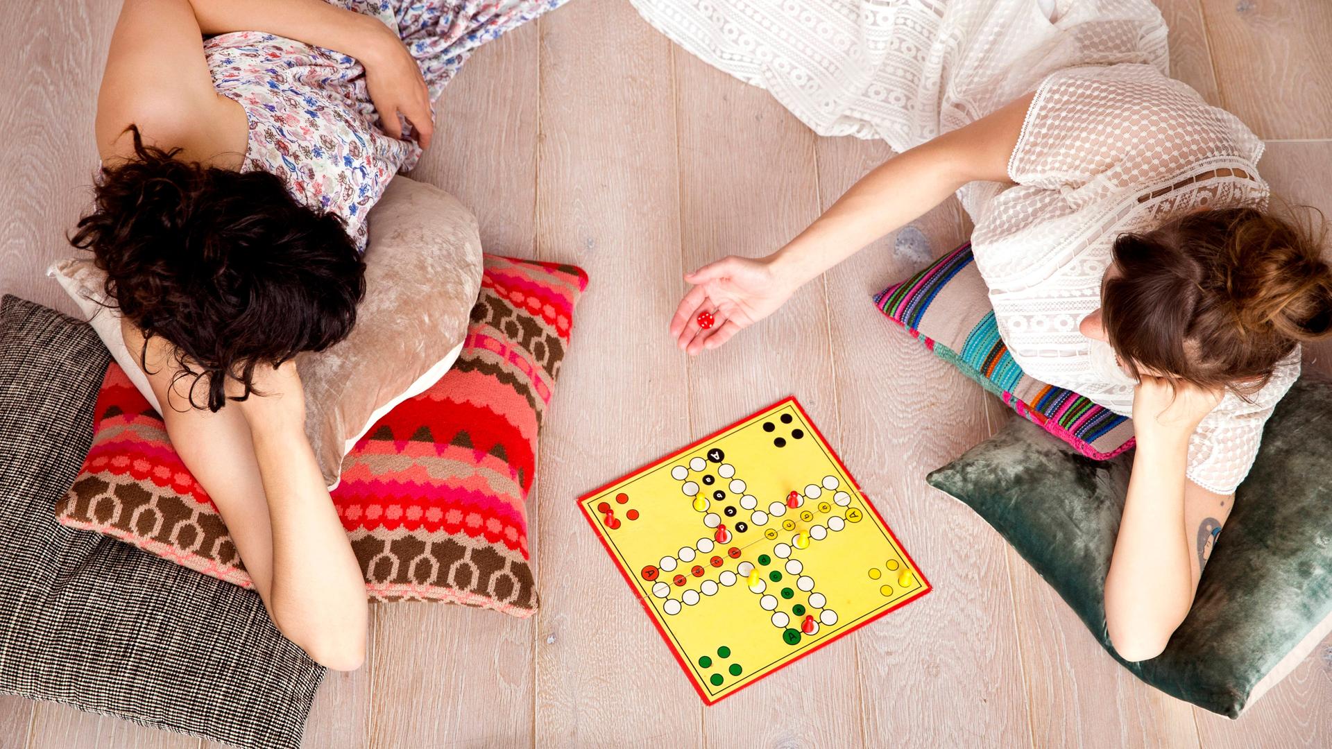 Zwei Frauen liegen auf bunten Kissen auf dem Fussboden und spielen Mensch-ärgere-dich-nicht.