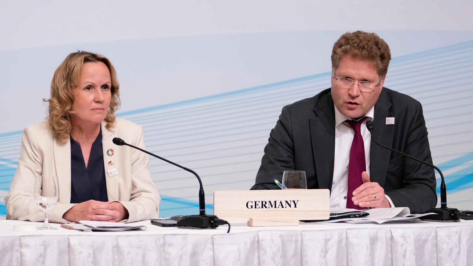 Die beiden sitzen nebeneinander an einem Tisch mit weißer Decke, vor ihnen Mikrofone. Graichen sagte etwas. Im Hintergrund eine weiße Wand mit einem blauen Netz aus Linien.