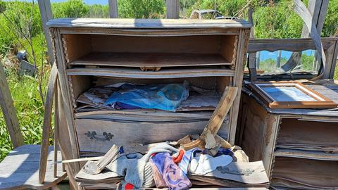 Eine alte Holzkommode steht in einer Hütte ohne Dach mitten in der Landschaft. Das Holz ist vom Regen aufgeweicht, in einer geöffneten Schublade sind noch persönliche Dinge der einstigen Bewohner zu sehen.