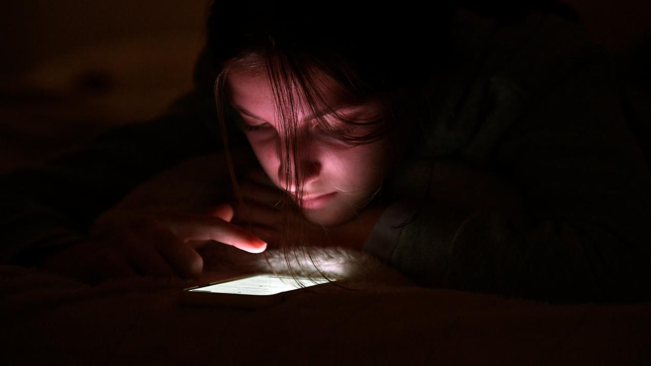 Eine Jugendliche nutzt ihr Handy bei Nacht in ihrem Zimmer. Der Finger scrollt über das leuchtende Display.