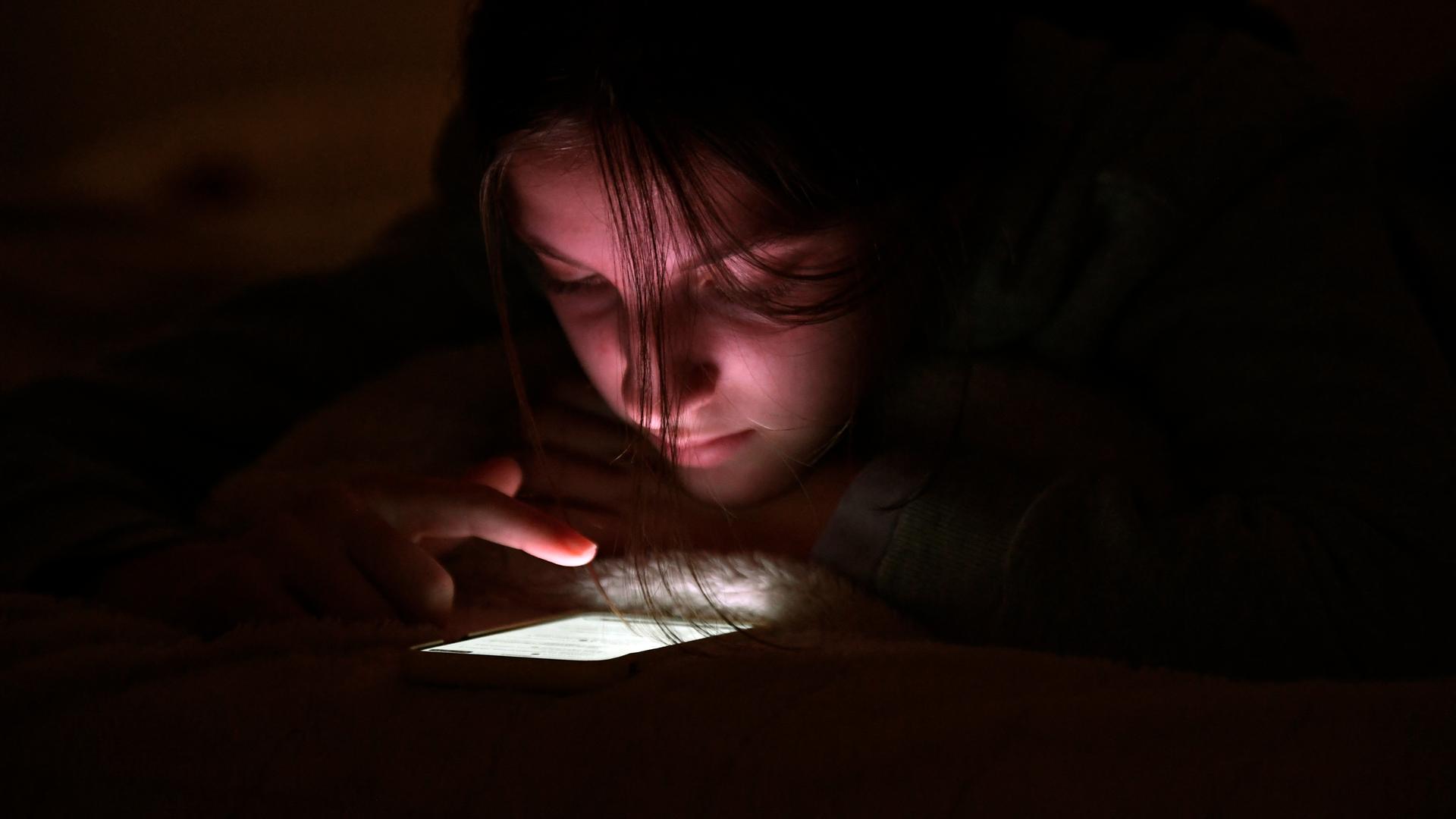 Eine Jugendliche nutzt ihr Handy bei Nacht in ihrem Zimmer. Der Finger scrollt über das leuchtende Display.