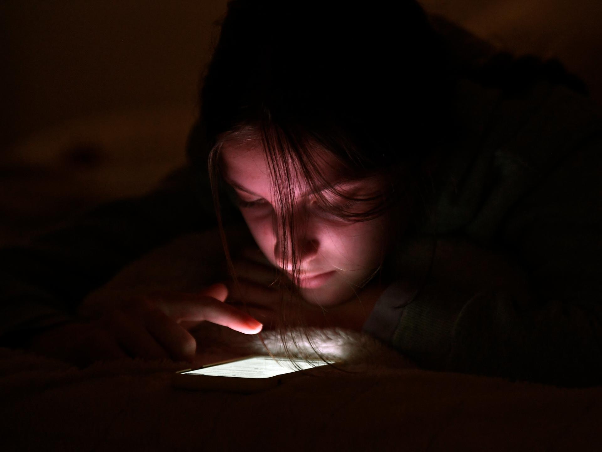 Eine Jugendliche nutzt ihr Handy bei Nacht in ihrem Zimmer. Der Finger scrollt über das leuchtende Display.