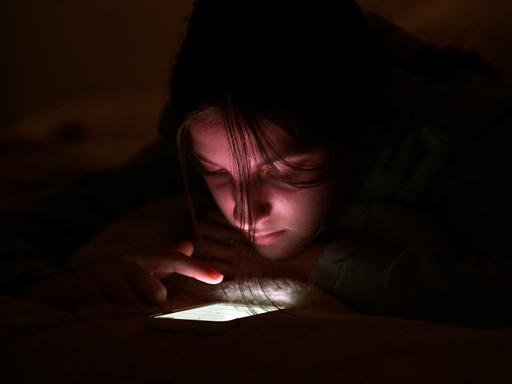 Eine Jugendliche nutzt ihr Handy bei Nacht in ihrem Zimmer. Der Finger scrollt über das leuchtende Display.