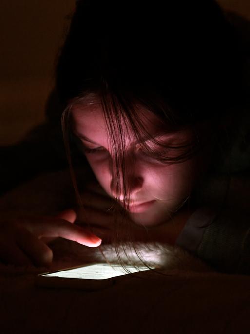 Eine Jugendliche nutzt ihr Handy bei Nacht in ihrem Zimmer. Der Finger scrollt über das leuchtende Display.