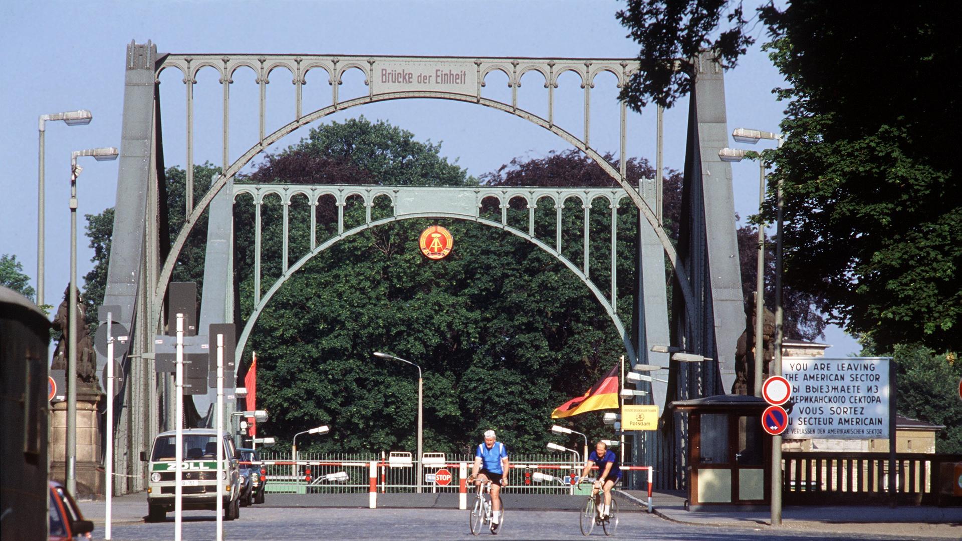 Die Glienicker Brücke an einem lichten Tag mit Fahrradfahrern. Im Hintergrund sind Bäume zu sehen.