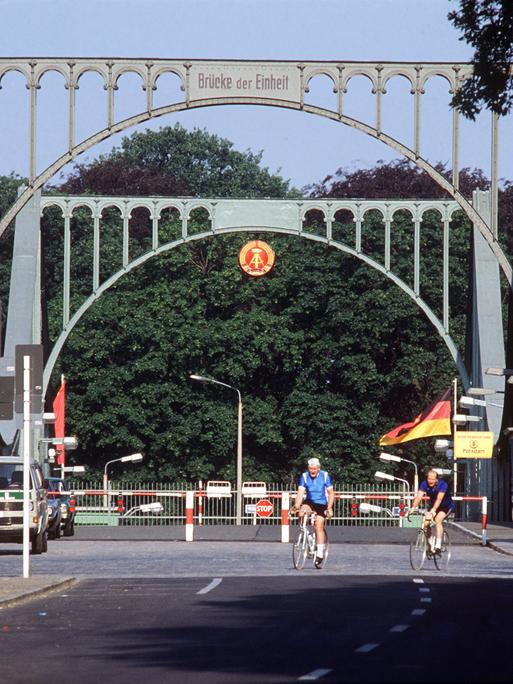 Die Glienicker Brücke an einem lichten Tag mit Fahrradfahrern. Im Hintergrund sind Bäume zu sehen.