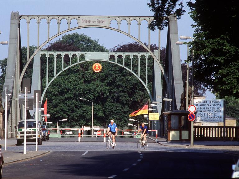 Die Glienicker Brücke an einem lichten Tag mit Fahrradfahrern. Im Hintergrund sind Bäume zu sehen.