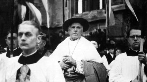 Schwarzweißfoto: Der katholische Theologe Clemens August Graf von Galen steht mit Kopfbedeckung und weißer Stola in einer Kirche und ist von anderen Menschen umgeben