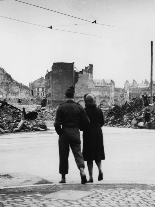 Ein US-Soldat geht mit einer deutscher Frau im Arm eine im Zweiten Weltkrieg zerstörte Straße entlang.