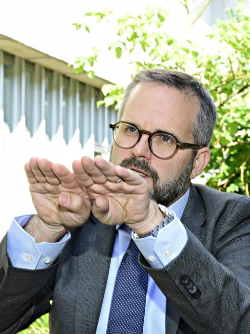 Matthias Berninger, Leiter des Bereiches Public and Governmental Affairs für die Bayer AG in, sitzt im Freien vor einem Gebäude. Im Hintergrund sind Gräser und grüne Gehölze zu sehen.