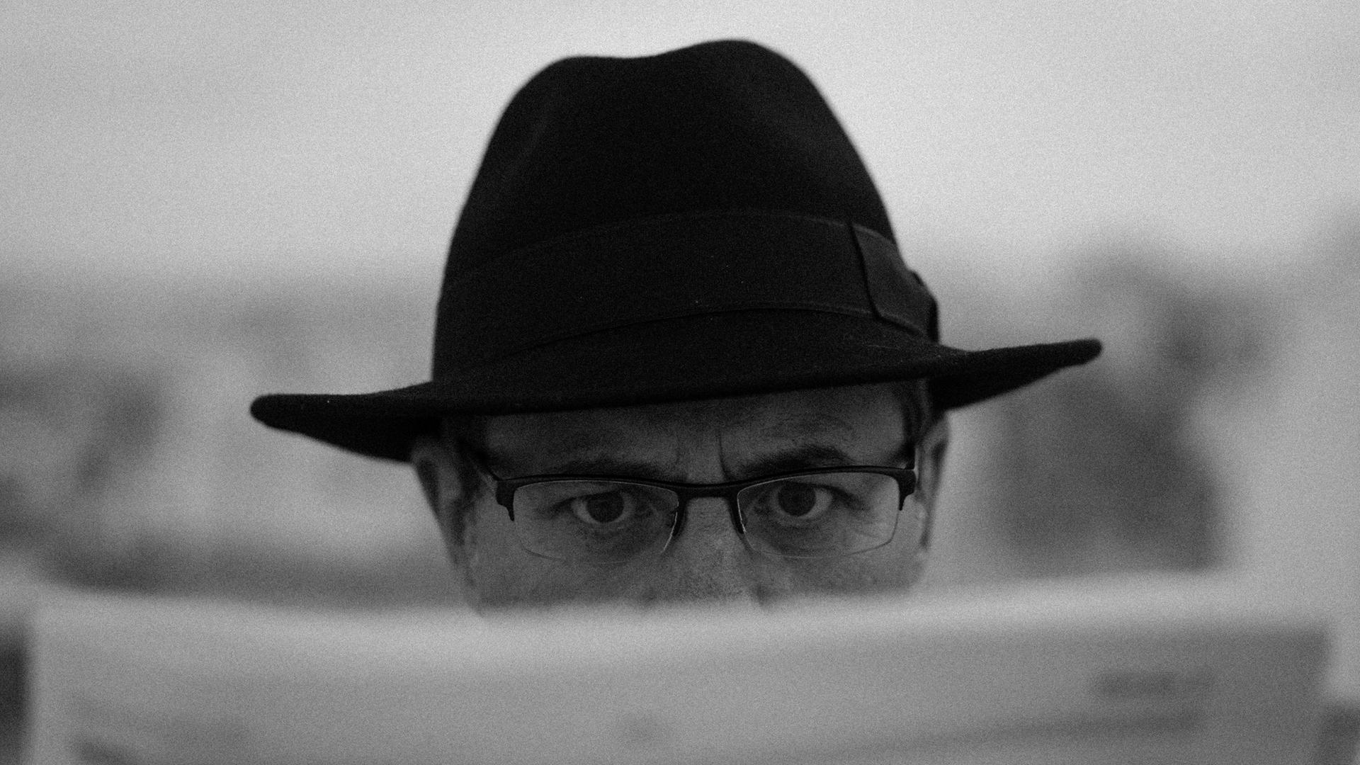 Schwarz-Weiß-Close-Up eines Manns mit Brille und Detektiv-Hut, der hinter einer Zeitung hervorblickt. Schwarz-Weiß-Close-Up eines Manns mit Brille und Detektiv-Hut, der hinter einer Zeitung hervorblickt.