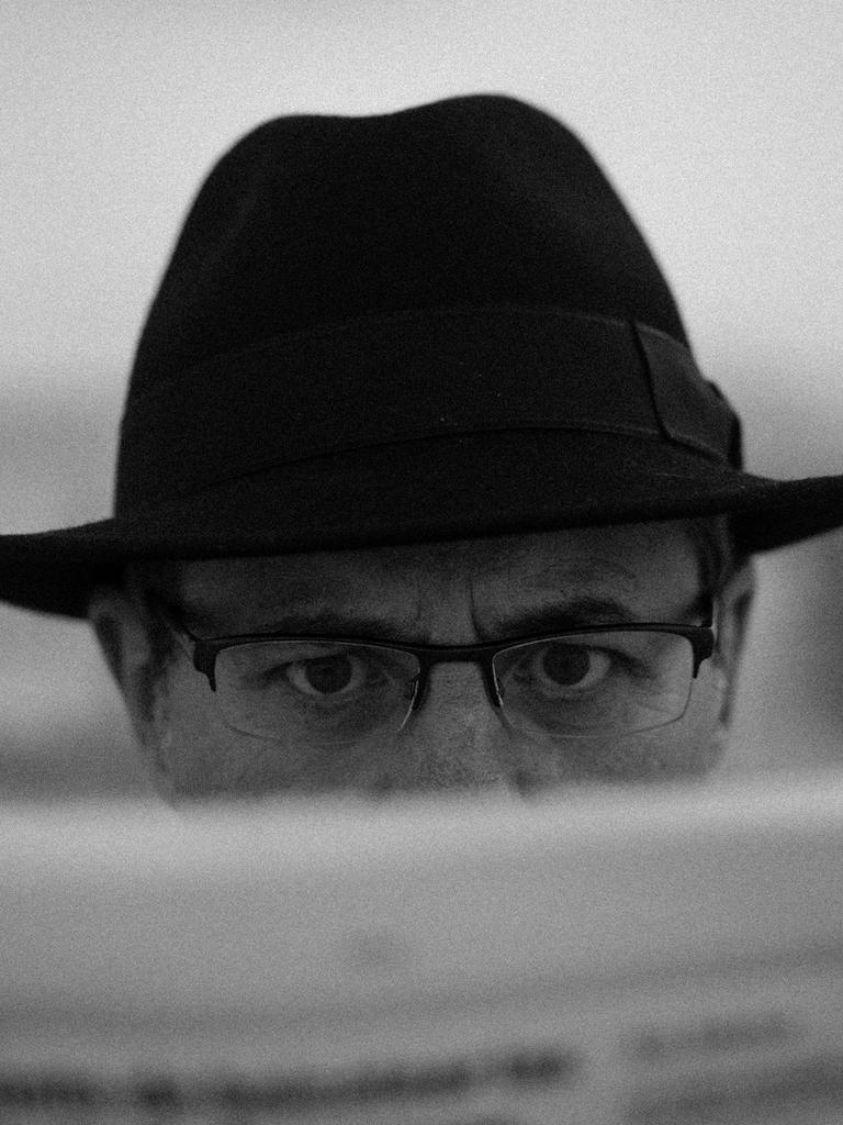 Schwarz-Weiß-Close-Up eines Manns mit Brille und Detektiv-Hut, der hinter einer Zeitung hervorblickt.