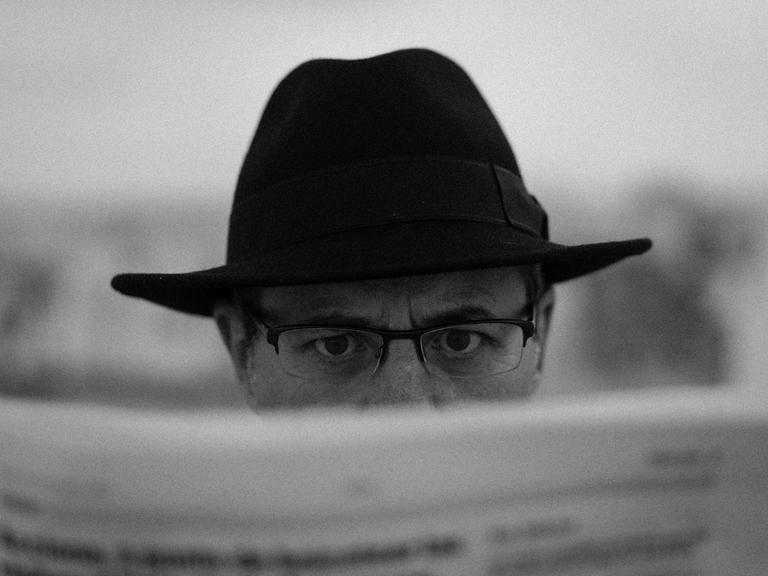 Schwarz-Weiß-Close-Up eines Manns mit Brille und Detektiv-Hut, der hinter einer Zeitung hervorblickt.