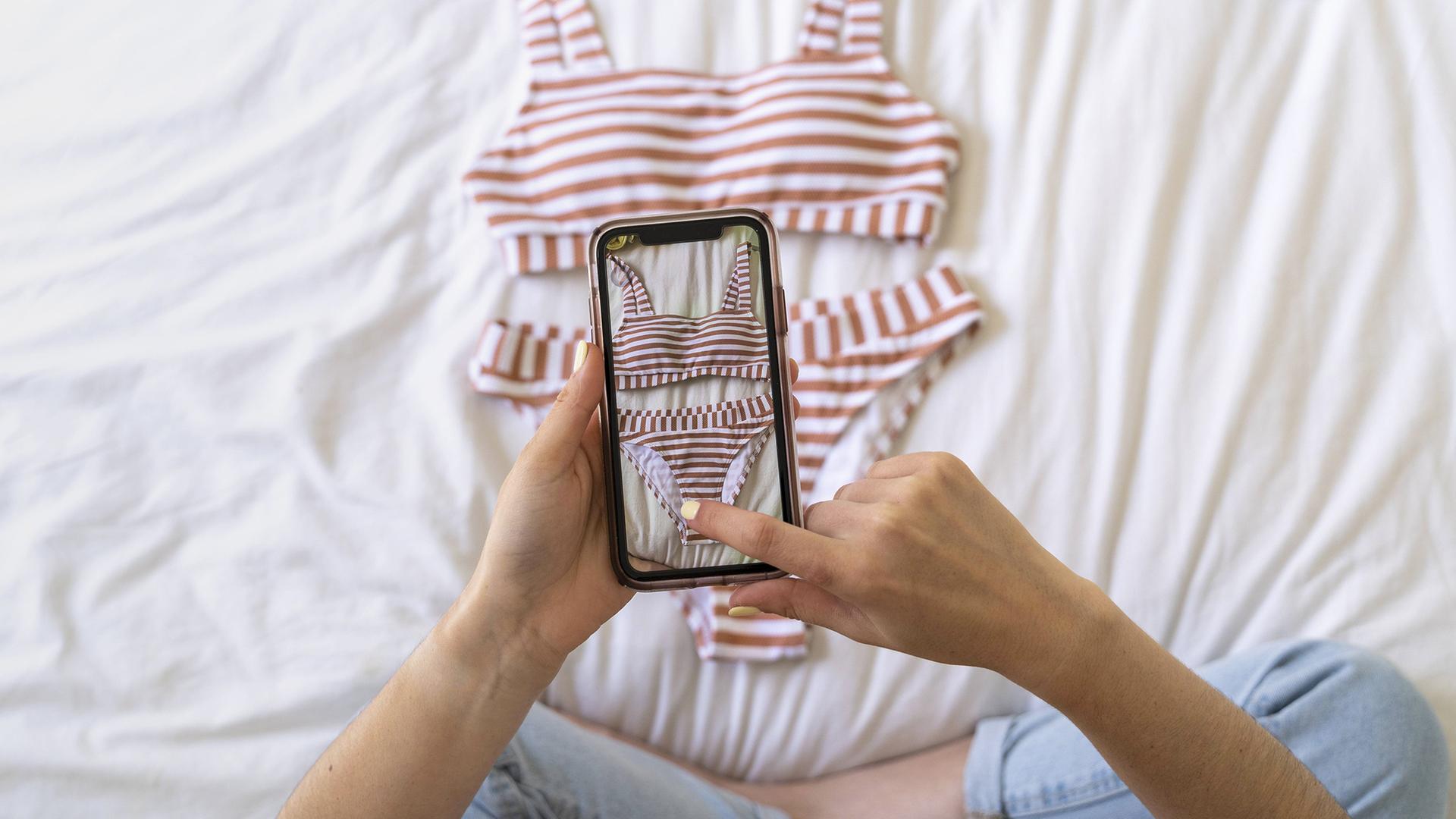 Eine junge Frau nimmt ein Smartphone-Foto von einem gestreiften Bikini auf ihrem Bett auf.