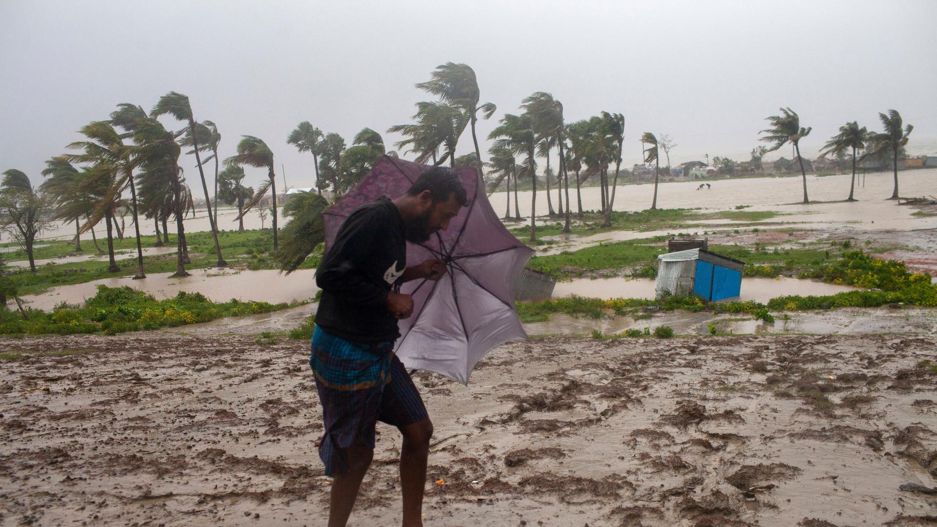 Ein Mann hält einen Schirm nach vorne, um sich vor Wind und Regen zu schützen. Im Hintergrund ist ein Küstenstreifen überflutet und Bäume wackeln im Wind. Ein Mann hält einen Schirm nach vorne, um sich vor Wind und Regen zu schützen. Im Hintergrund ist ein Küstenstreifen überflutet und Bäume wackeln im Wind.