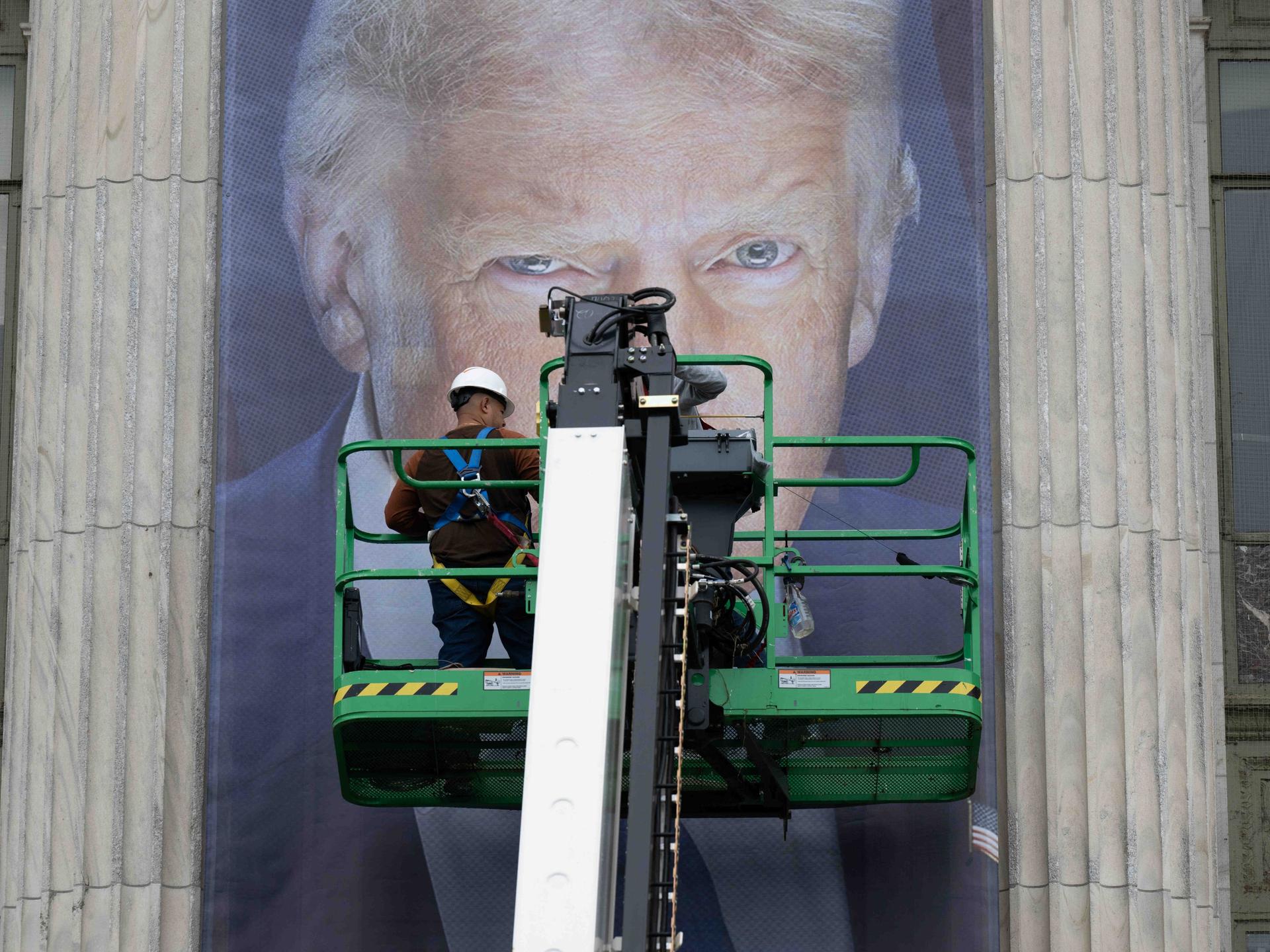 Donald Trump als Plakat. Seine Augen sind hinter einer Plattform zu erkennen, auf der ein Arbeiter steht. Vermutlich bringt dieser gerade das überlebensgroße Banner an