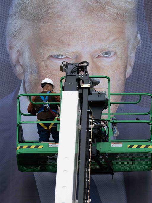 Donald Trump als Plakat. Seine Augen sind hinter einer Plattform zu erkennen, auf der ein Arbeiter steht. Vermutlich bringt dieser gerade das überlebensgroße Banner an