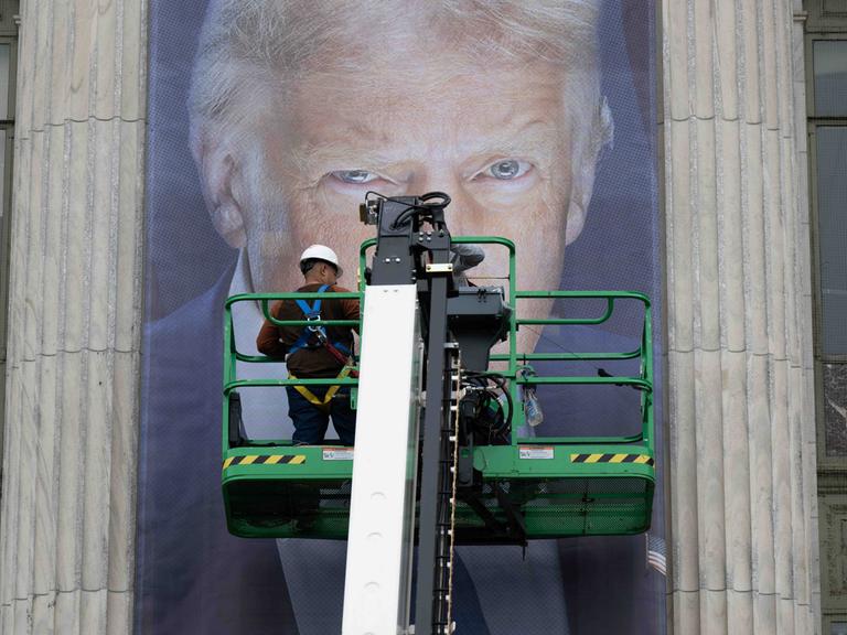 Donald Trump als Plakat. Seine Augen sind hinter einer Plattform zu erkennen, auf der ein Arbeiter steht. Vermutlich bringt dieser gerade das überlebensgroße Banner an