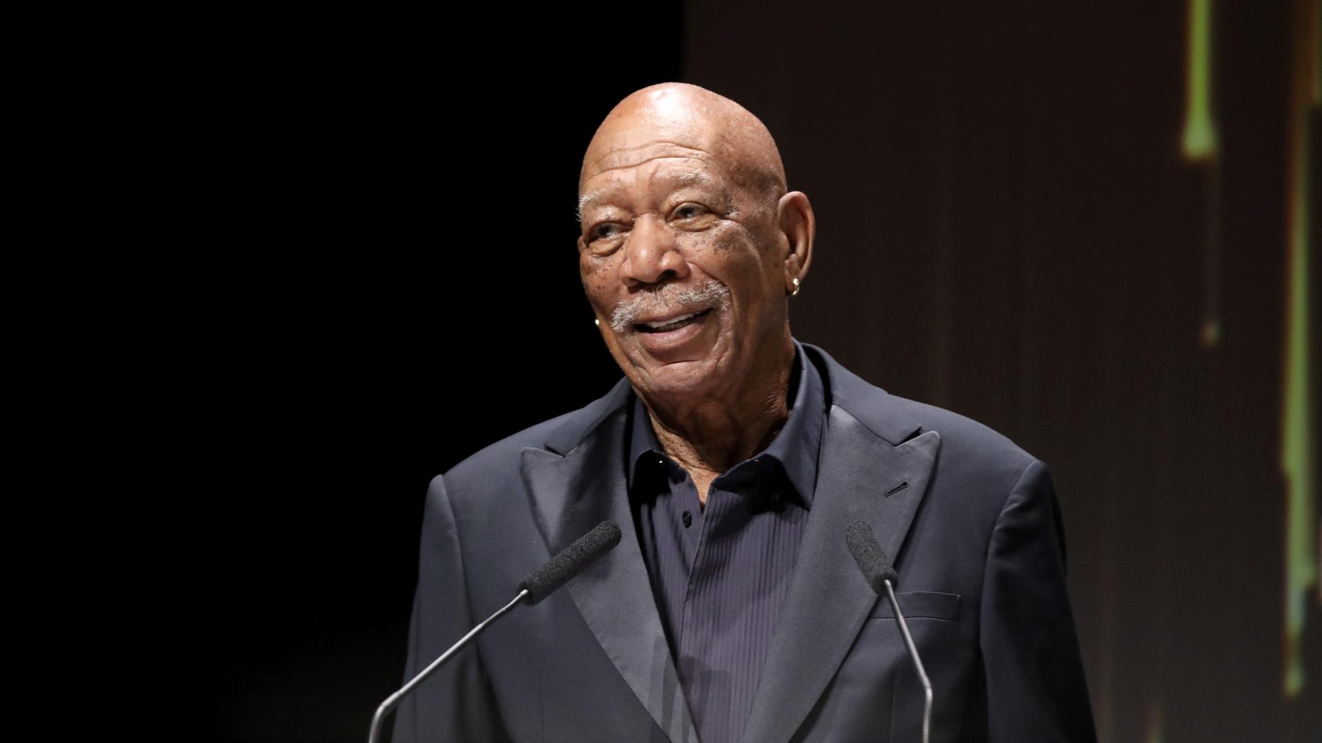 Morgan Freeman steht an einem Rednerpult und lächelt. Er hat einen weißen Schnurrbart und eine Glatze. Morgan Freeman steht an einem Rednerpult und lächelt. Er hat einen weißen Schnurrbart und eine Glatze.