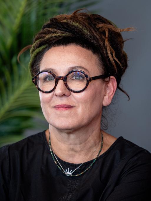 Olga Tokarczuk trägt eine runde, schwarz umrandete Brille und ein schwarzes Oberteil. Sie schaut zur Seite und lächelt.