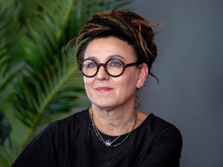 Olga Tokarczuk trägt eine runde, schwarz umrandete Brille und ein schwarzes Oberteil. Sie schaut zur Seite und lächelt.