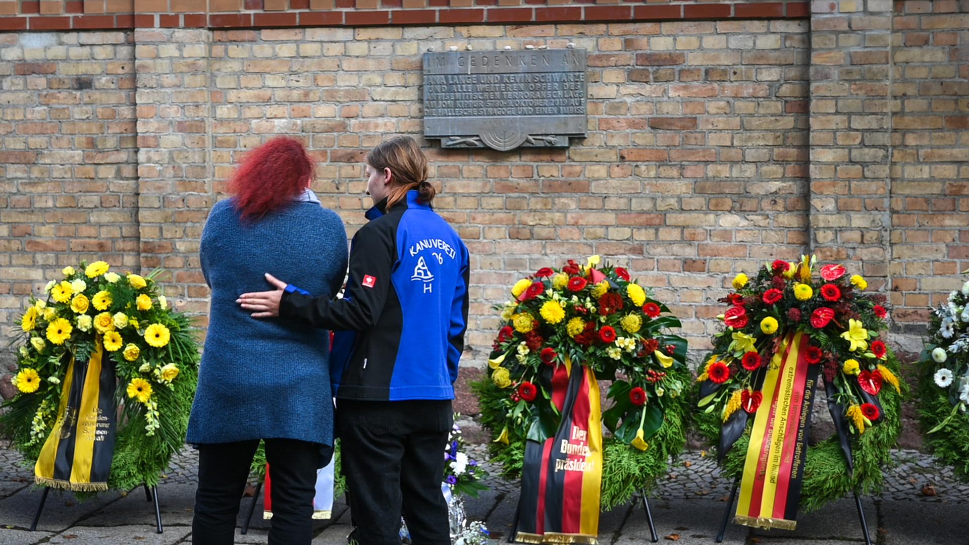 Zwei Frauen stehen vor vier Kränzen. Eine Frau legt ihre Hand auf den Rücken von der anderen Frau. Die Kränze liegen vor einer Mauer. Sie erinnern an den Anschlag von Halle.