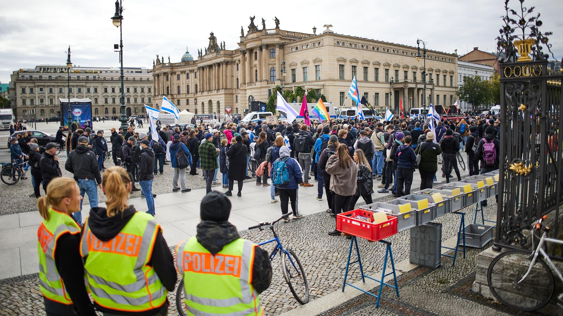 Eine pro-israelische Demonstration vor der Humboldt-Universität in Berlin. Im Vordergrund stehen drei Menschen mit gelben Westen mit der Aufschrift "Polizei".