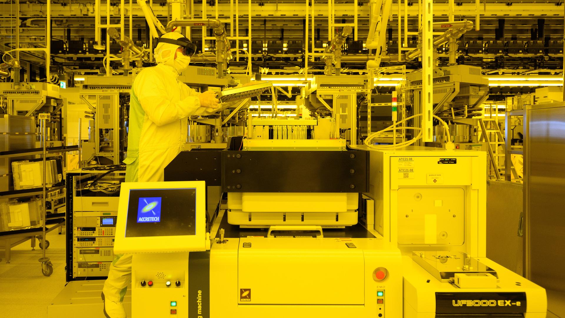 Ein Mitarbeiter im Ganzkörperschutzanzug demonstriert Wartungsarbeiten an einer Prüfmaschine für Chips mit Augmented-Reality-Brille.