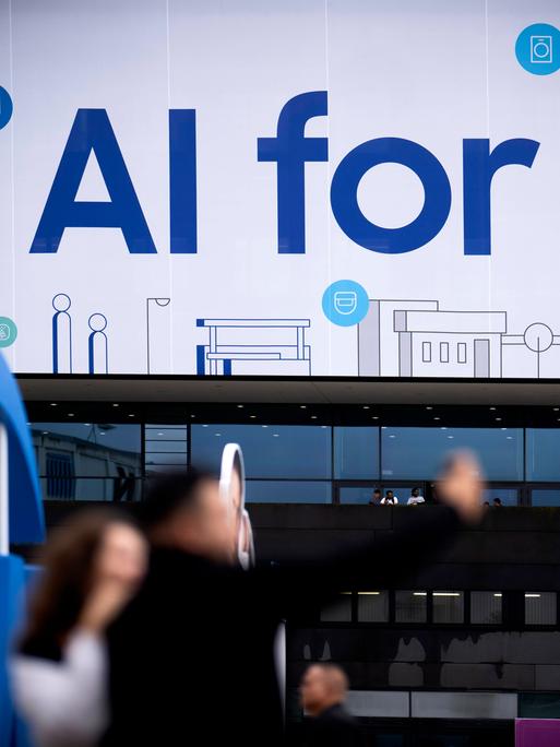 An einem Samsung Stand auf der IFA Berlin im Jahr 2024 steht in großen Buchstaben "KI for all" geschrieben. Davor sind in der Unschärfe einige Messebesucher zu sehen.