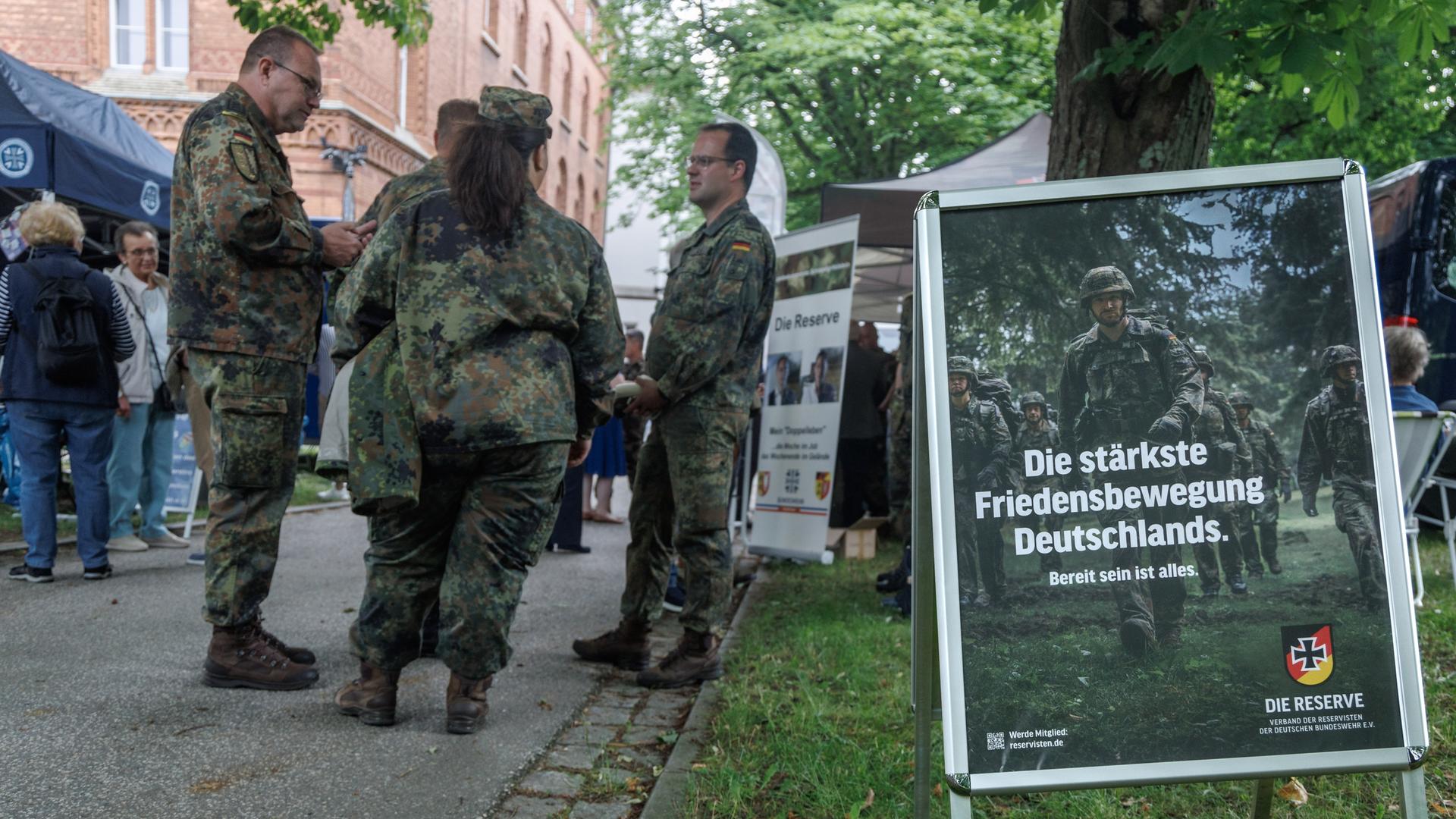 Informationsstände auf dem Veteranentag in Kiel. Bundeswehrsoldaten stehen in einer Gruppe. Daneben ist ein Plakat aufgestellt mit der Aufschrift: Die stärkste Friedensbewegung Deutschlands. 