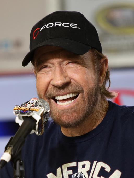 Der Schauspieler Chuck Norris sitzt vor einem Mikrofon und lacht.