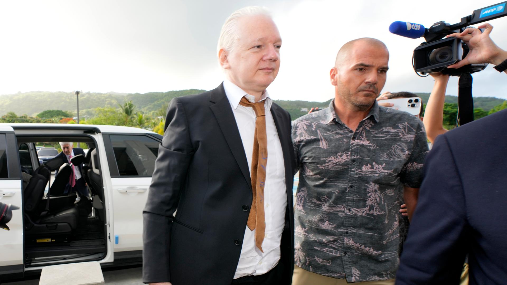 Assange mit schwarzem Anzug und brauner Krawatte wird von einem Mann im Haiwaii-Hemd eskortiert. Dahinter haltne zwei Personen eine kamera und ein handy hoch. Im Hintergrund das Auto, mit dem Assange eintraf.
