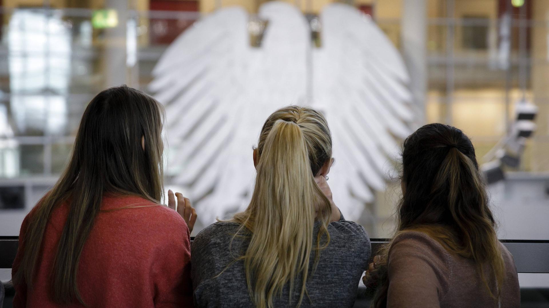 Drei junge Frauen einer Besuchergruppe blicken auf den Plenarsaal im Bundestag. Im Hintergrund ist der Bundestagsadler zu sehen, das Wappentier des Plenarsaals.