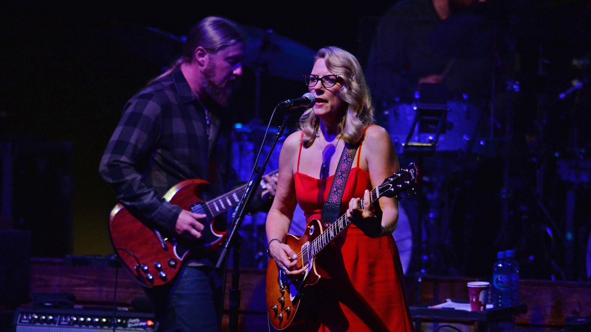 Eine Frau in rotem Kleid und Gitarre singt vor einem Mikrofon. Ein Mann mit Gitarre und Vollbart steht hinter ihr und spielt ebenso.