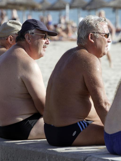 Vier ältere Männer sitzen an einem Strand auf Mallorca auf einer Mauer und blicken aufs Wasser.