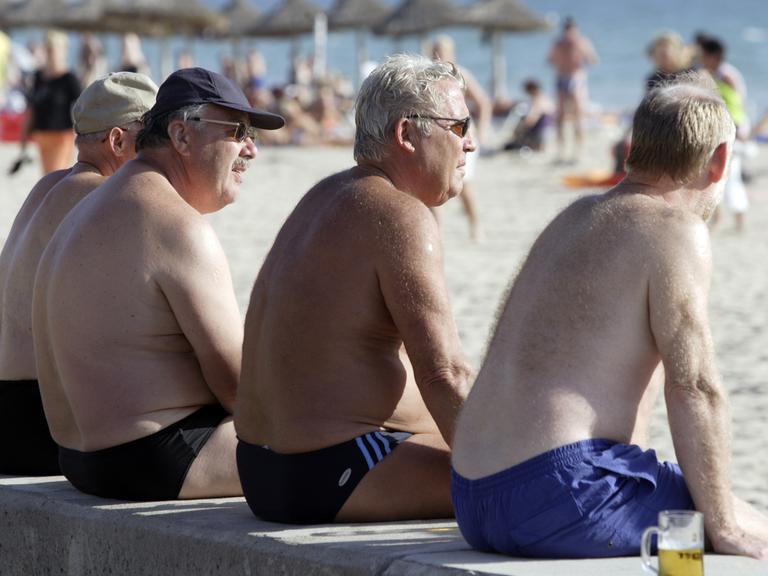 Vier ältere Männer sitzen an einem Strand auf Mallorca auf einer Mauer und blicken aufs Wasser.