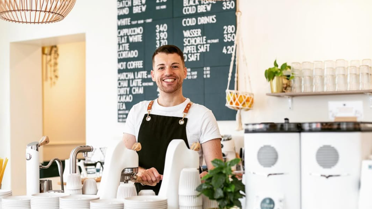 Hinter dem Tresen steht ein lächelnder Mann mit Schürze, der in die Kamera blickt.
