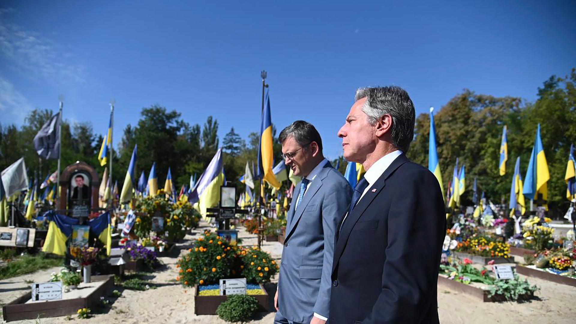 Zwei Männer in Anzügen stehen auf einem Soldatenfriedhof, im Hintergrund sind Gräber mit ukrainischen Flaggen und Blumen zu sehen.