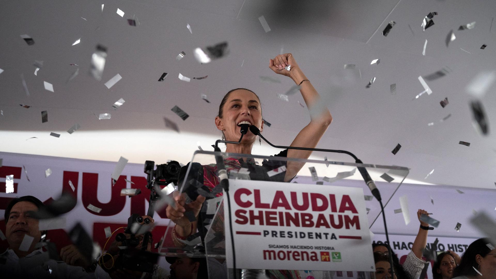 Claudia Sheinbaum steht auf einem Podium und reckt die rechte Faust in die Höhe, während Konfetti auf sie herabregnet.