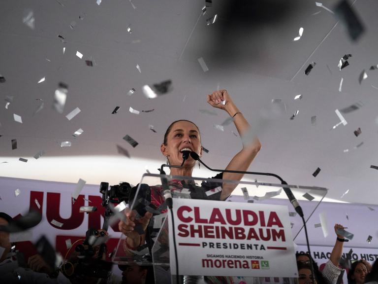 Claudia Sheinbaum steht auf einem Podium und reckt die rechte Faust in die Höhe, während Konfetti auf sie herabregnet.